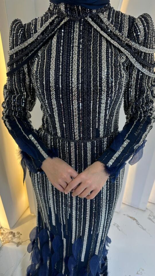 Elbisenin gövdesi boyunca uzanan dikey çizgisel pullu ve boncuklu işlemeler Omuzlardan sarkan inci veya boncuk zincirlerden Yüksek yakalı ve uzun kollu Lacivert Abiye 44