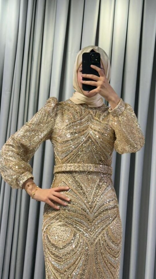 Altın rengi boncuklar, pullar ve payetlerle yoğun bir şekilde işlenmiş,   Vücuda oturan, uzun bir silüete Yüksek yakalıdır ve omuzları hafifçe vatkalı, balon kesim, uzun şeffaf tül kollara Gold Abiye 46