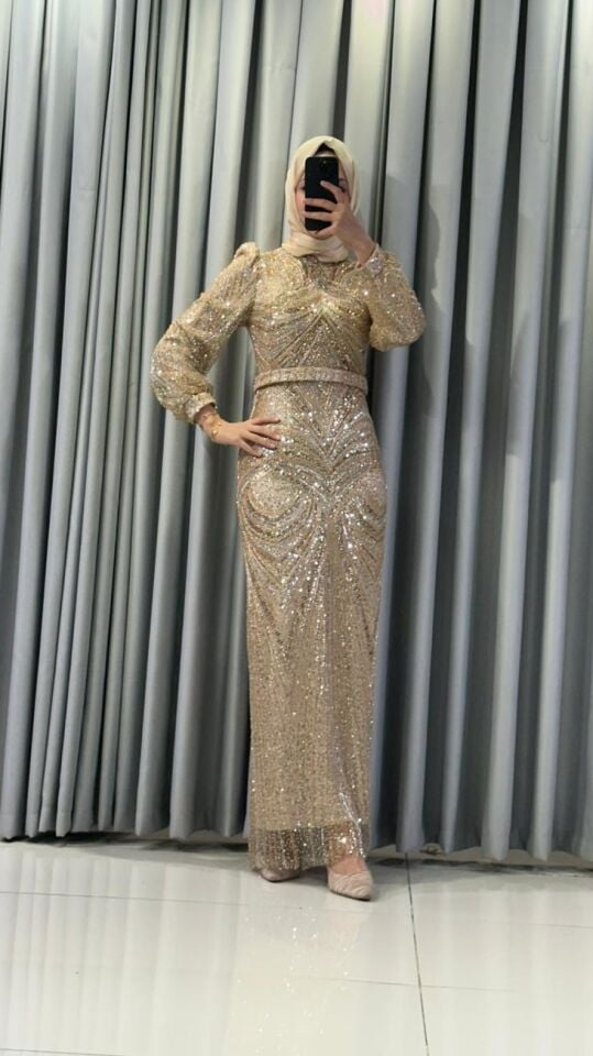 Altın rengi boncuklar, pullar ve payetlerle yoğun bir şekilde işlenmiş,   Vücuda oturan, uzun bir silüete Yüksek yakalıdır ve omuzları hafifçe vatkalı, balon kesim, uzun şeffaf tül kollara Gold Abiye 46