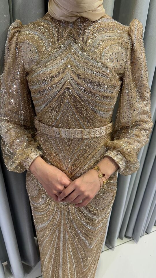 Altın rengi boncuklar, pullar ve payetlerle yoğun bir şekilde işlenmiş,   Vücuda oturan, uzun bir silüete Yüksek yakalıdır ve omuzları hafifçe vatkalı, balon kesim, uzun şeffaf tül kollara Gold Abiye 46