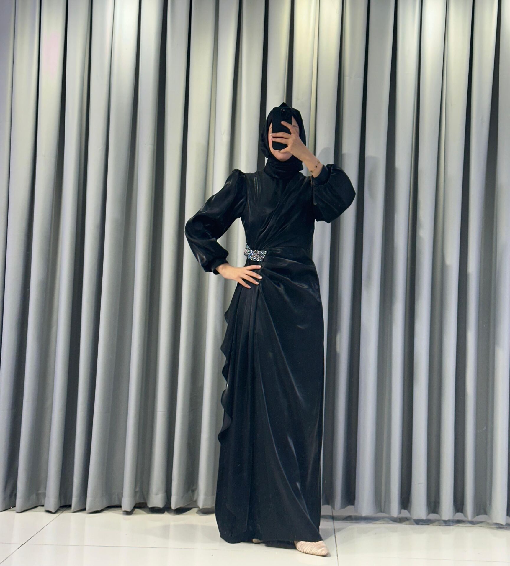 Uzun kollu, saten kumaştan yapılmış omuzları kabarık/vatkalı ve belinde taşlı, dekoratif bir broş detayına sahiptir Eteği, önden drapeli ve katmanlı bir tasarımla yere kadar uzanmaktadır (Siyah Abiye) 44