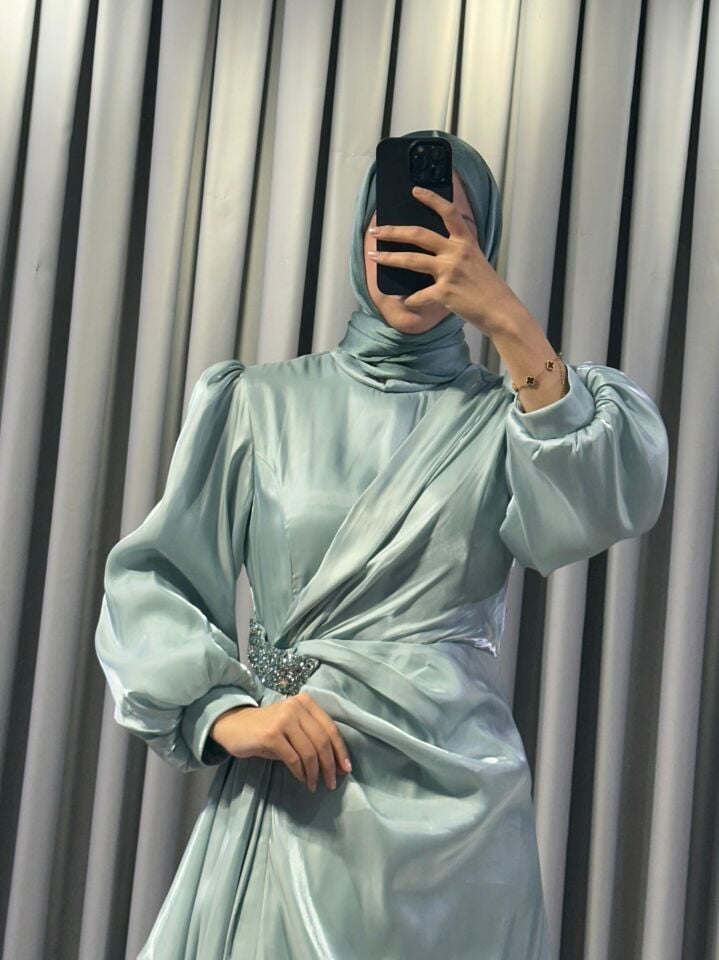 Uzun kollu, saten kumaştan yapılmış omuzları kabarık/vatkalı ve belinde taşlı, dekoratif bir broş detayına sahiptir Eteği, önden drapeli ve katmanlı bir tasarımla yere kadar uzanmaktadır ( Mind Yeşili  Abiye) 44