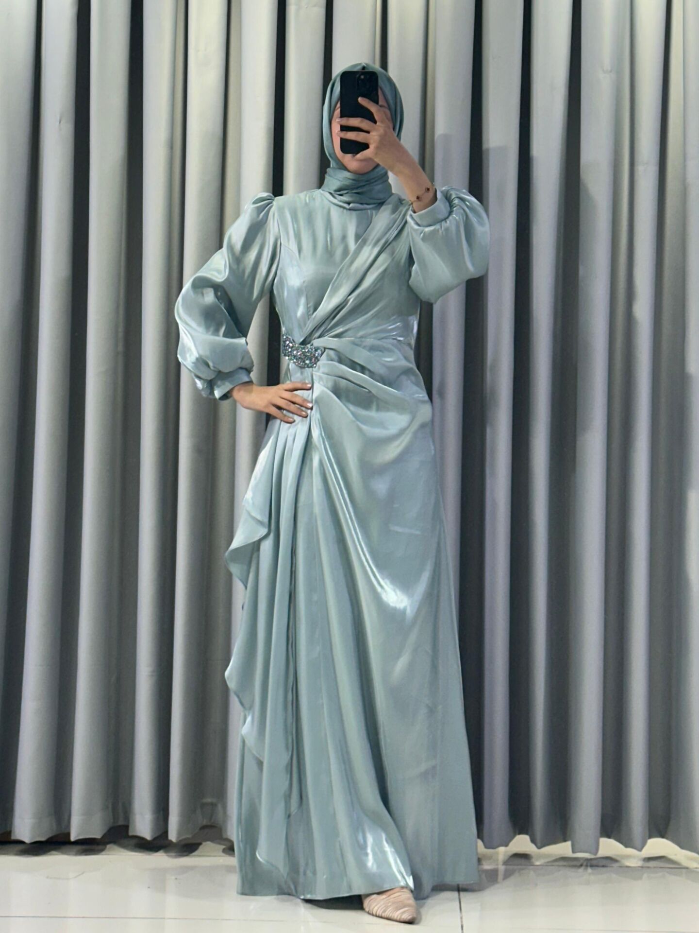 Uzun kollu, saten kumaştan yapılmış omuzları kabarık/vatkalı ve belinde taşlı, dekoratif bir broş detayına sahiptir Eteği, önden drapeli ve katmanlı bir tasarımla yere kadar uzanmaktadır ( Mind Yeşili  Abiye) 38